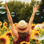 Woman wearing a straw hat and red top standing in a vibrant sunflower field, arms raised joyfully toward the sky, surrounded by floating bubbles — symbolizing freedom, happiness, and the rejuvenating power of self-care.
