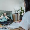 A coach and client communicate through a video call on a laptop. The coach on the screen is smiling and giving two thumbs up, while the client sits at a desk with notebooks, a laptop, and books nearby, symbolizing an online coaching session and positive connection.
