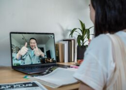 A coach and client communicate through a video call on a laptop. The coach on the screen is smiling and giving two thumbs up, while the client sits at a desk with notebooks, a laptop, and books nearby, symbolizing an online coaching session and positive connection.