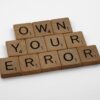 Scrabble tiles spelling “Own Your Error” on a wooden surface, symbolizing personal responsibility, growth, and the power of accountability in coaching.
