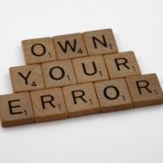 Scrabble tiles spelling “Own Your Error” on a wooden surface, symbolizing personal responsibility, growth, and the power of accountability in coaching.