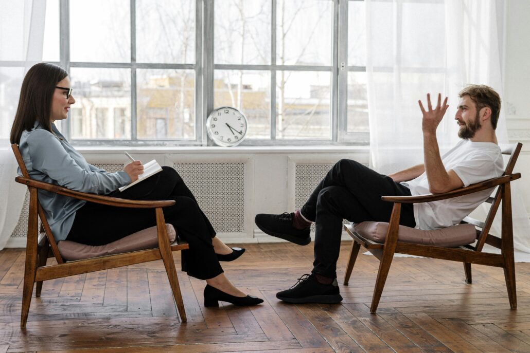 A life coach and client sit facing each other in a bright, minimalist office with wooden floors and large windows. The coach, holding a notebook, listens attentively as the client gestures while speaking, illustrating a focused one-on-one coaching session.