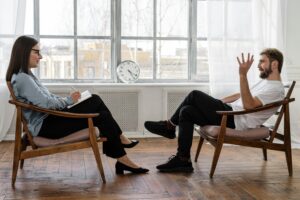 A life coach and client sit facing each other in a bright, minimalist office with wooden floors and large windows. The coach, holding a notebook, listens attentively as the client gestures while speaking, illustrating a focused one-on-one coaching session.