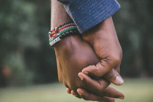 Relationship Coaching Certification: Building Stronger Connections – A close-up of two people holding hands, symbolizing trust, support, and emotional connection. One person wears a beaded bracelet with vibrant colors, adding a touch of cultural significance. The blurred background suggests an outdoor setting, emphasizing warmth and togetherness.