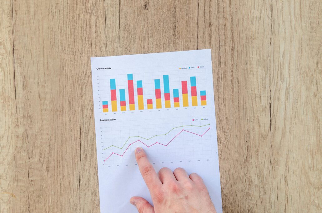 A hand points to a printed financial report showing colorful bar and line graphs representing company performance metrics like sales, receipts, and orders—symbolizing the measurable ROI of life coaching and personal growth.