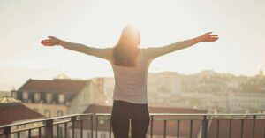A woman stands on a balcony with her arms outstretched, facing the morning sun over a city skyline. The warm sunlight creates a glowing, hopeful atmosphere, symbolizing confidence, freedom, and self-empowerment — aligning with the theme of overcoming imposter syndrome and embracing authenticity.