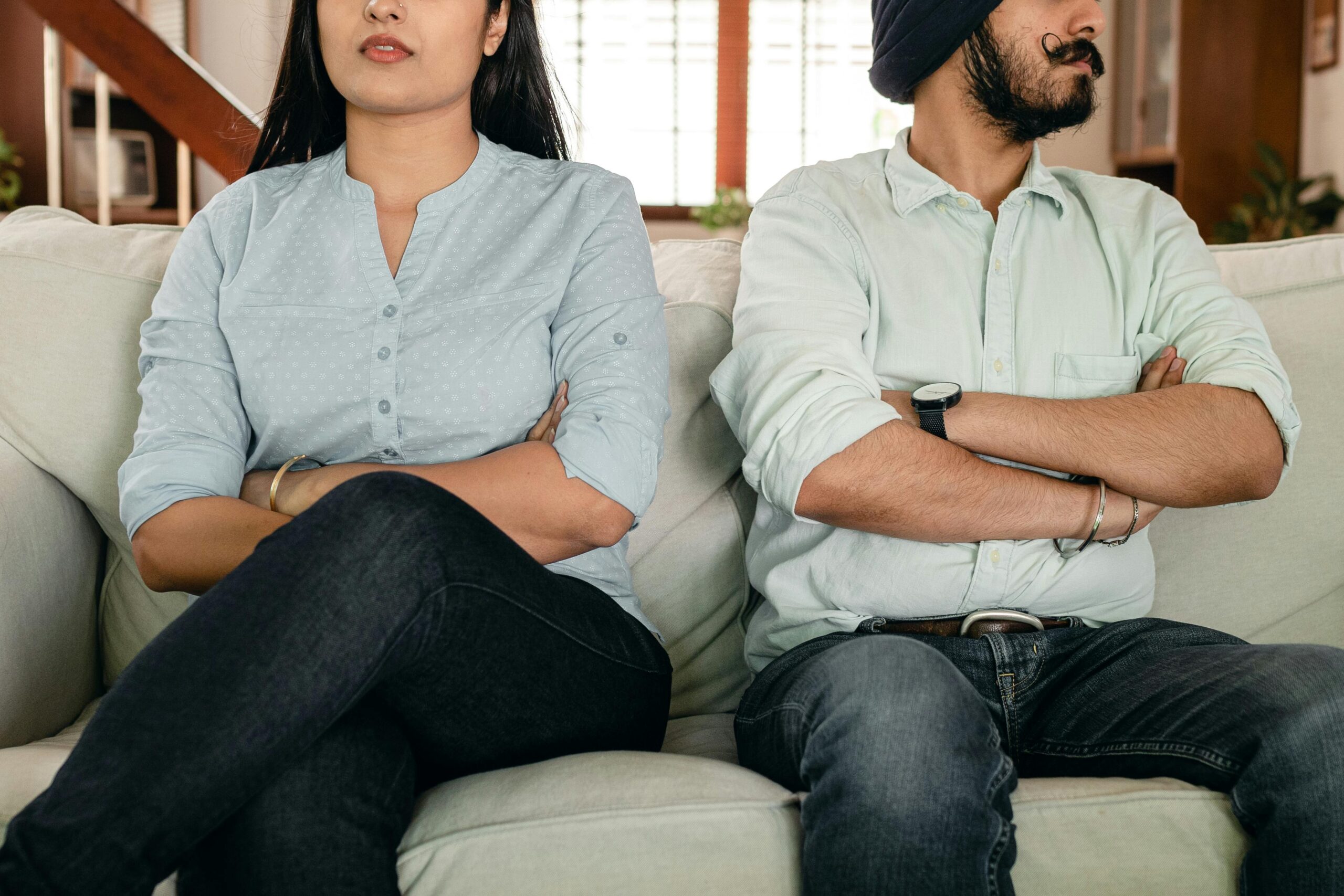 Couple sitting on a couch with arms crossed, showing tension during a difficult conversation, illustrating conflict resolution coaching.