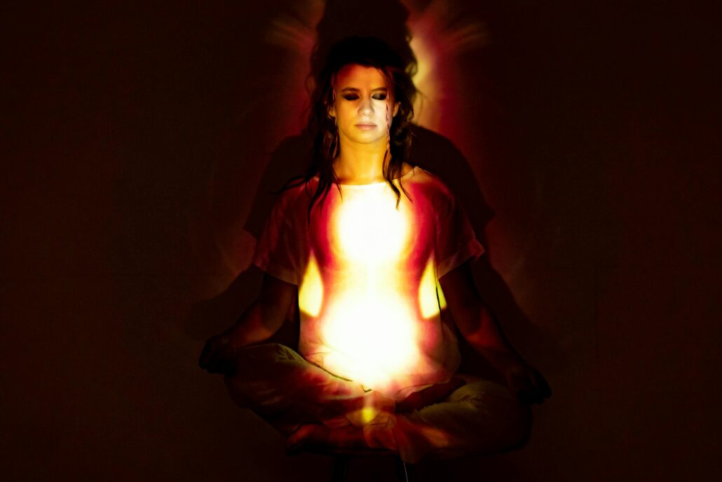A person sitting cross-legged in a meditative posture with eyes closed, illuminated by warm red and yellow light patterns that create a glowing, hypnotic effect across their chest and face.