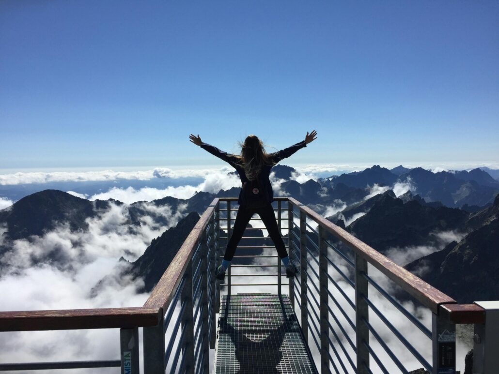 Person standing with arms wide open on a mountain viewpoint above the clouds, symbolizing freedom, possibility, and The Power of AND™.