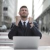 A determined man in a business suit sitting at a laptop outdoors, clenching his fists in a moment of emotional resilience or triumph, symbolizing overcoming setbacks through inner strength.