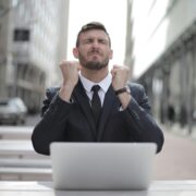 A determined man in a business suit sitting at a laptop outdoors, clenching his fists in a moment of emotional resilience or triumph, symbolizing overcoming setbacks through inner strength.