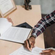 A person signing documents on a wooden desk with a certificate and pen holder in the background, symbolizing professionalism and certification for Life coach certification