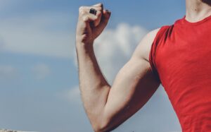 A close-up of a certified health coach's flexed arm, showcasing toned muscles. The person is wearing a red sleeveless shirt and a black ring on their finger. The background features a blue sky with white clouds.